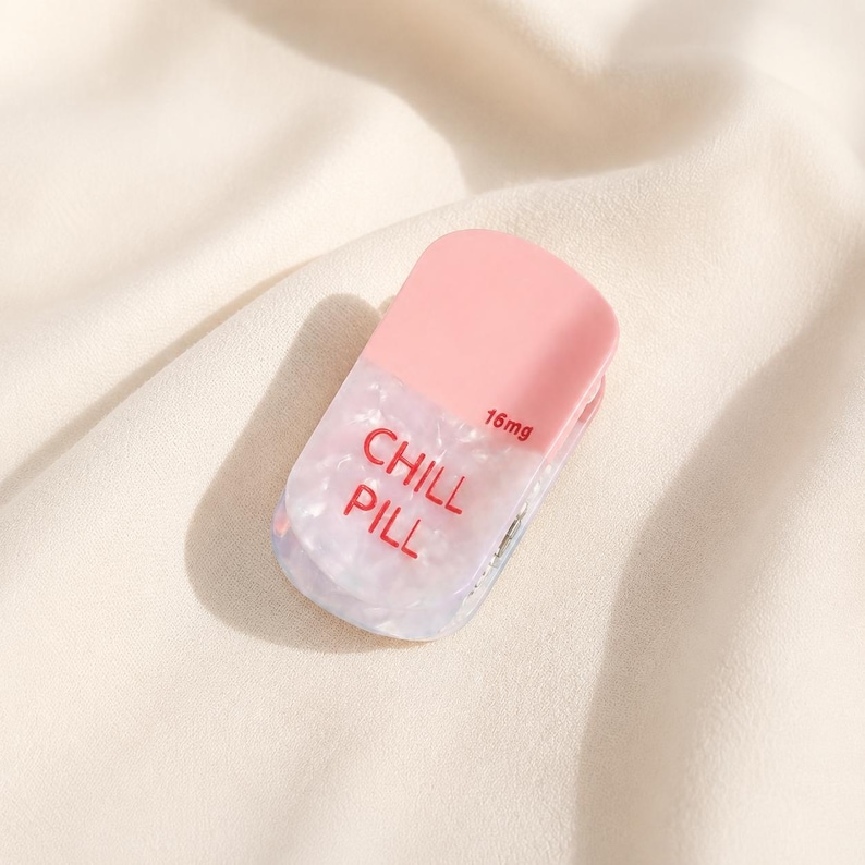 May include: A pill-shaped hair clip featuring a solid light pink top section and a translucent, pearlescent white bottom section. The text CHILL PILL is printed in red on the bottom, with the text 16mg printed in smaller red font above it.