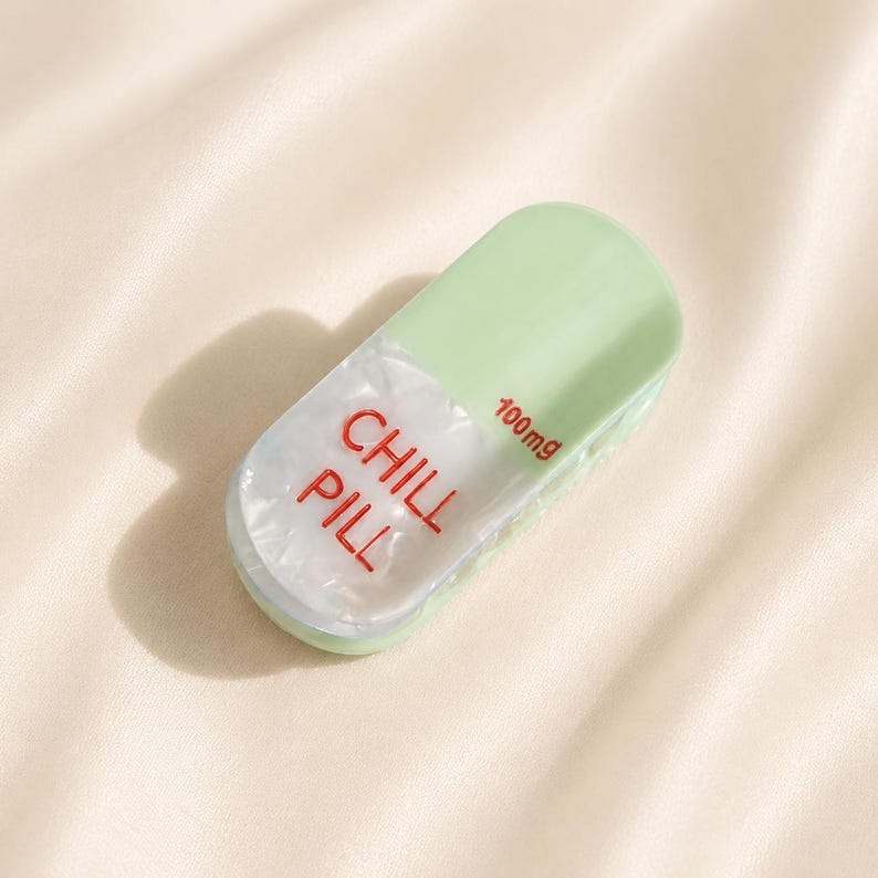 May include: A capsule-shaped object featuring a light green top and a white, pearlescent bottom section. The text CHILL PILL is printed in red on the white portion, with the text 100mg also in red near the top edge of the white section.