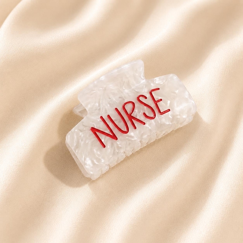 May include: A white hair claw clip with a marbled texture and the word NURSE written in bold red capital letters across the front. The accessory features a rectangular shape with a spring-loaded mechanism and interlocking teeth for securing hair.