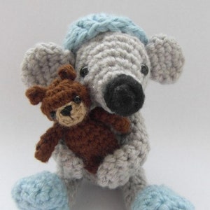 Peut inclure: Une souris en crochet gris portant un chapeau bleu tient un ours en peluche en crochet marron. La souris a des chaussons en crochet bleus.