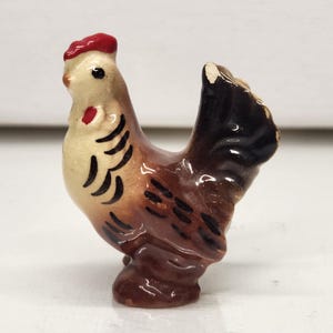 May include: A small ceramic rooster figurine. The rooster has a red comb, a cream-colored body, and brown and black markings. The tail is black and brown. The figurine is standing on a brown base.