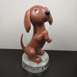 May include: A brown dachshund figurine with green eyes and a black nose stands on its hind legs. The dog is perched on a round, marble-patterned base with the text "I W". The figurine has large, floppy ears and a curved tail.