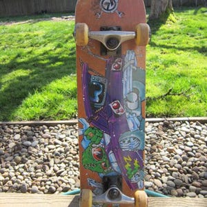 May include: A skateboard with a brown deck featuring a colorful graphic of cartoon characters and shapes. The board has yellow wheels, silver trucks, and a logo near the top. The skateboard is designed for recreational use.