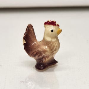 May include: A small ceramic figurine of a chicken. The chicken has a brown body, a white chest, and a red comb. It has a yellow beak and black eyes. The figurine is standing on a white surface.