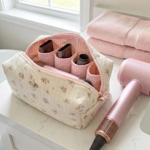 Peut inclure: Trousse de maquillage à imprimé floral avec intérieur rose à carreaux, contenant des outils de coiffure. Un sèche-cheveux rose avec une buse cuivrée est posé à côté du sac, avec deux serviettes roses en arrière-plan.