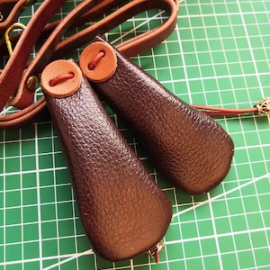 Puede incluir: Primer plano de dos empuñaduras de correa para perros de cuero marrón oscuro con un detalle de botón. Las empuñaduras están sobre una superficie de cuadrícula verde, con una correa de cuero marrón a juego en el fondo.