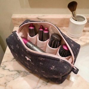 May include: A dark gray quilted cosmetic bag with pink floral accents, open to reveal hair styling tools. The bag has a pink zipper and interior lining. A white ceramic holder with a makeup brush is to the right.