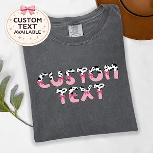 May include: A charcoal gray t-shirt featuring the words CUSTOM TEXT in a bold font. The letters have a black and white cow print pattern on the top half and a pink diagonal striped pattern on the bottom half, accented with small pink bows.