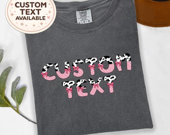May include: A charcoal gray t-shirt featuring the words CUSTOM TEXT in a bold font. The letters have a black and white cow print pattern on the top half and a pink diagonal striped pattern on the bottom half, accented with small pink bows.