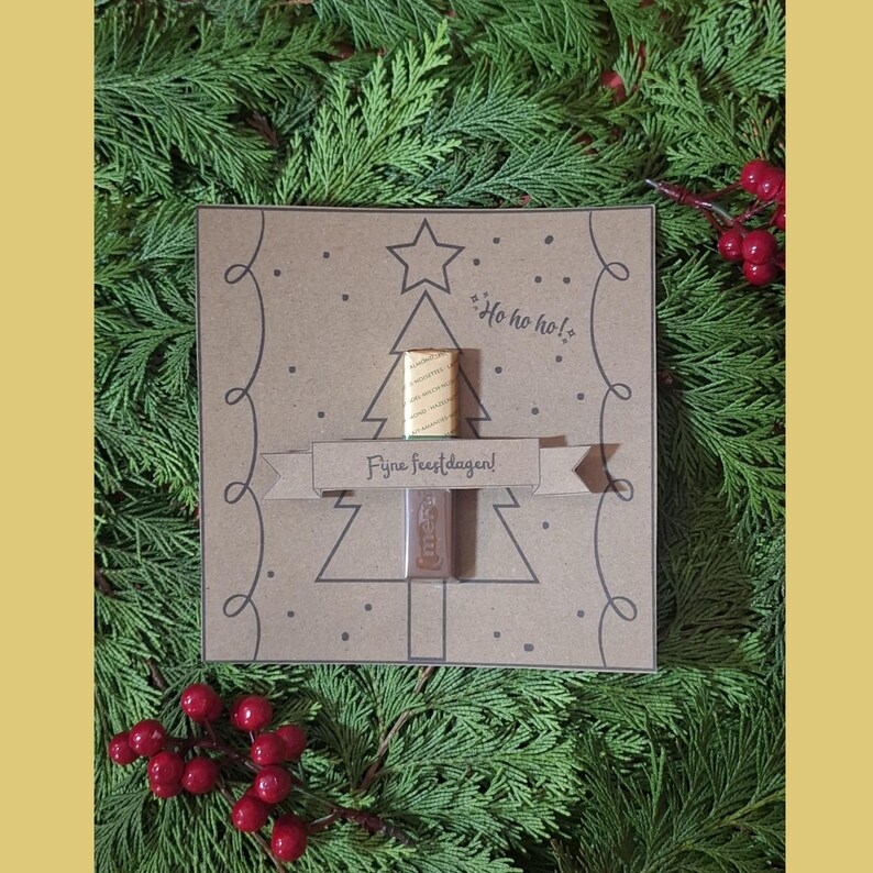 May include: A festive Christmas card with a tree design, featuring the phrases "Ho ho ho!" and "Fijne feestdagen!". The card holds two small items. The image is set against a backdrop of green foliage and red berries.