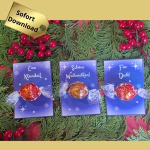 Op de afbeelding: Drie kleine blauwe kaarten met sneeuwvlokontwerpen en Duitse tekst, elk met een verpakte chocolade. De kaarten liggen op een bed van groen met rode bessen en een gouden label met "Sofort Download".