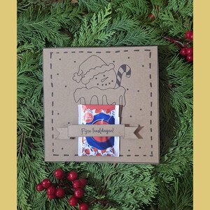 May include: A brown square gift box featuring a snowman illustration and a candy cane. A small packet with a red, white, and blue design is attached. The text "Fijne feestdagen!" is on a banner. The background is evergreen branches and red berries.