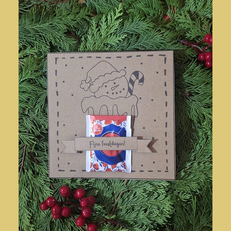 May include: A brown square gift box featuring a snowman illustration and a candy cane. A small packet with a red, white, and blue design is attached. The text "Fijne feestdagen!" is on a banner. The background is evergreen branches and red berries.