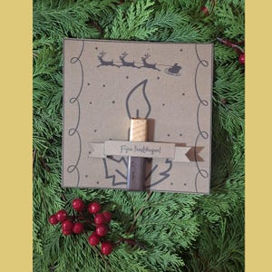 May include: A brown Christmas card featuring a candle design, the text "Fijne feestdagen!", and a drawing of Santa's sleigh. The card is decorated with red berries and set on a bed of green foliage.