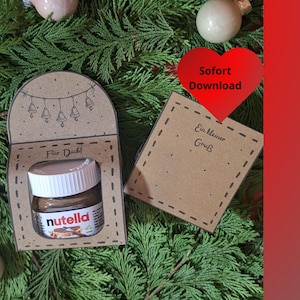 Peut inclure: Un petit pot de Nutella dans une boîte cadeau en papier brun, avec les mots "Für Dich!" imprimés sur le devant. Une petite carte avec les mots "Ein kleiner Gruß" est à côté de la boîte. Un cœur rouge avec les mots "Sofort Download" est également visible.