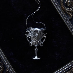 May include: A silver-colored pendant necklace featuring a detailed, ornate chalice design. The chalice has a fluted rim, decorative handles, and floral embellishments. The pendant hangs from a delicate silver chain against a dark background.