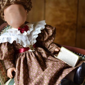 Op de afbeelding: Een bruine en witte gebloemde jurk met een witte kanten kraag en een rood lint. De jurk wordt gedragen door een pop met bruin haar en een boek in haar hand.