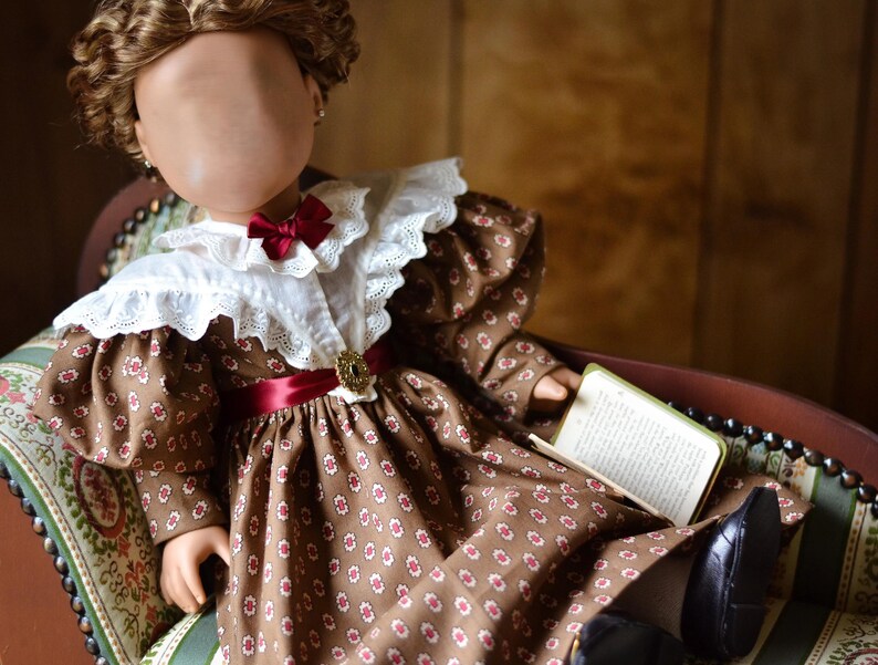 Op de afbeelding: Een bruine en witte gebloemde jurk met een witte kanten kraag en een rood lint. De jurk wordt gedragen door een pop met bruin haar en een boek in haar hand.