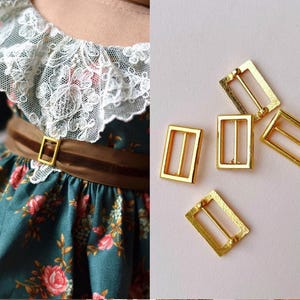 Puede incluir: Un vestido floral verde azulado con cuello de encaje blanco y cinturón marrón con hebilla dorada. También se muestran varias hebillas rectangulares doradas, adecuadas para proyectos de costura o artesanía. El vestido presenta rosas rosas y rojas.