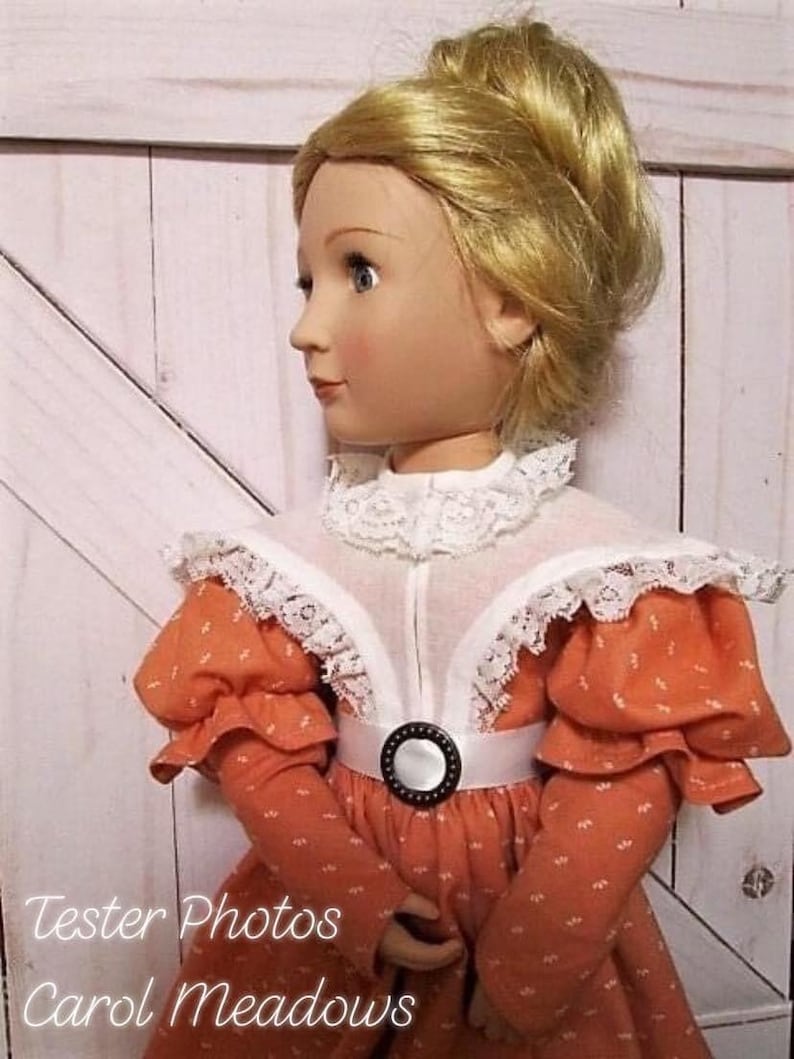 Op de afbeelding: Een pop die een perzikkleurige jurk draagt met witte kantversiering en een zwart-witte riem. De pop heeft blond haar dat in een knotje is gemodelleerd. De pop staat voor een witte houten deur.