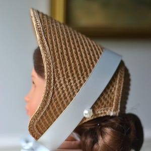 May include: A doll wearing a vintage-style bonnet. The bonnet is made of woven straw with a light blue ribbon and a faux pearl accent. The doll has brown hair and is wearing a pink dress.
