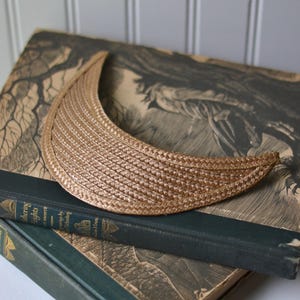 May include: A woven, crescent-shaped sun visor in a light tan color. The visor is resting on top of two antique books with dark green covers and gold lettering. The background is a light gray, vertical-paneled wall.