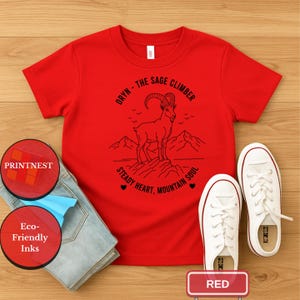 May include: A red t-shirt with a black graphic of a mountain goat and the text "ORYN - THE SAGE CLIMBER STEADY HEART, MOUNTAIN SOUL." The shirt is paired with light blue jeans and white sneakers.