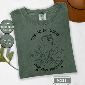 May include: A moss green t-shirt with a graphic of a mountain goat and the text "ORYN - THE SAGE CLIMBER." The shirt also features the text "STEADY HEART, MOUNTAIN SOUL." The shirt is made of 100% ring-spun cotton and is part of the Comfort Colors collection.