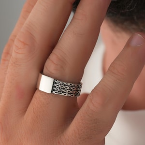 Op de afbeelding: Een zilveren ring met een rechthoekige bovenkant en een kettingontwerp rond de band. De ring wordt om een vinger gedragen en toont de ingewikkelde details en de metalen glans. De ring is een sieraad.