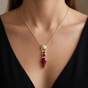 May include: A gold necklace with a pendant featuring a large, red, gemstone at the bottom, two smaller red gemstones, and a diamond-studded top. The necklace is worn against a black background.