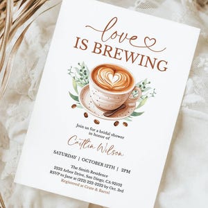 K&ouml;nnte beinhalten: Eine wei&szlig;e Einladungskarte mit dem Text "love IS BREWING" und einer Kaffeetassen-Illustration. Die Karte ist f&uuml;r eine Brautparty zu Ehren von Caitlin Wilson, mit Datum, Uhrzeit und Ortsangaben. Die Karte liegt auf einem wei&szlig;en Spitzentuch.