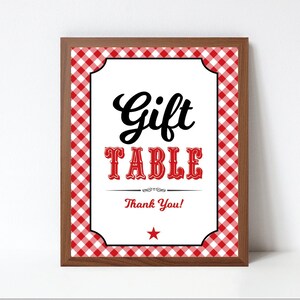 Op de afbeelding: Afdrukbaar bord met een rode en witte gingham achtergrond. Het bord zegt "Gift Table" in zwarte tekst met een rode ster eronder. Het bord is ingelijst in bruin hout.