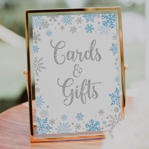 Puede incluir: Un cartel con marco dorado, fondo blanco y diseños de copos de nieve azules y plateados. El cartel dice "Tarjetas y Regalos".