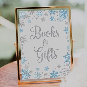 May include: A gold-framed sign with the words "Books & Gifts" in silver glitter script. The sign is decorated with blue and silver snowflakes. The sign is on a wooden surface.