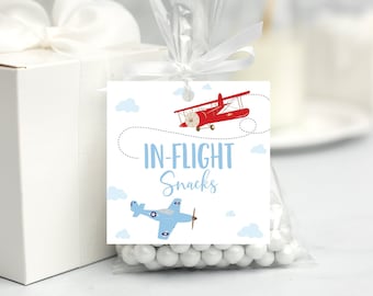 May include: A white gift box with a white bow sits beside a clear bag of white sweets. The bag has a tag with a red biplane and a blue biplane, with the words "IN-FLIGHT Snacks" in blue.