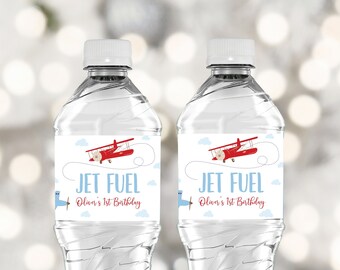 May include: Two clear plastic water bottles with blue and white labels. The labels feature a red airplane with white clouds and the text "Jet Fuel Oliver's 1st Birthday".