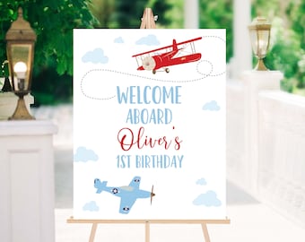 May include: Welcome aboard Oliver's 1st birthday sign featuring a red and blue aeroplane illustration. The sign is on a wooden easel.
