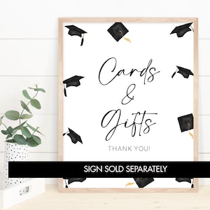 May include: A white sign with black text that reads "Cards & Gifts Thank You!"  The sign is framed in a light wood frame.  The background is a white wood panel.  There are black graduation caps with gold tassels scattered around the sign.