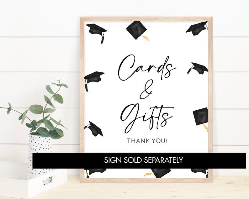 May include: A white sign with black text that reads "Cards & Gifts Thank You!"  The sign is framed in a light wood frame.  The background is a white wood panel.  There are black graduation caps with gold tassels scattered around the sign.