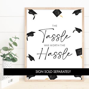 May include: A white graduation sign with black text that reads "The Tassle Was Worth The Hassle". The sign is decorated with black graduation caps with gold tassels. The text "Sign Sold Separately" is at the bottom of the sign.