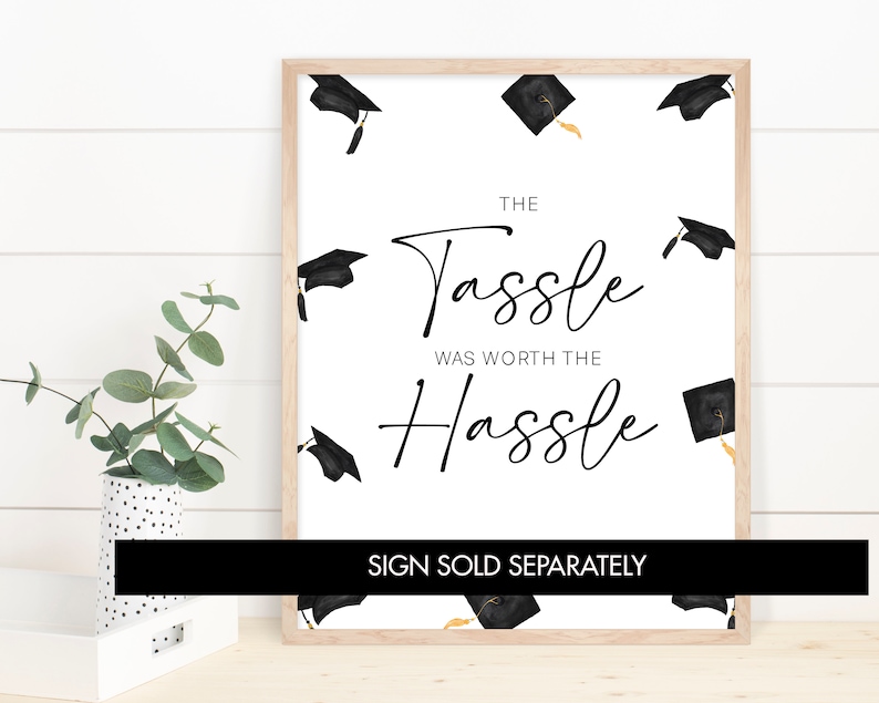 May include: A white graduation sign with black text that reads "The Tassle Was Worth The Hassle". The sign is decorated with black graduation caps with gold tassels. The text "Sign Sold Separately" is at the bottom of the sign.