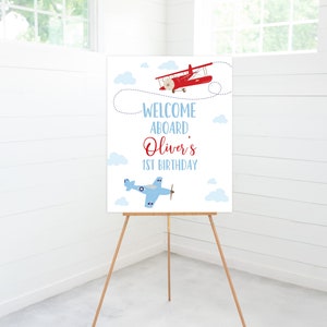 May include: Welcome aboard Oliver's 1st birthday sign with a red and white airplane and a blue and white airplane on a white background with blue clouds.