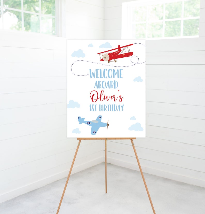 May include: Welcome aboard Oliver's 1st birthday sign with a red and white airplane and a blue and white airplane on a white background with blue clouds.