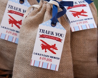 May include: Thank you for flying by favor bags with a red airplane design and blue and white striped border. The bags are made of burlap and have a blue ribbon tied around them. The text on the tags reads "Thank you for flying by" and "Laura's Baby Shower! 11-09-2015".
