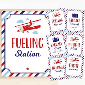 Puede incluir: Señales de fiesta con temática de avión rojo y azul con texto que dice "Estación de combustible", "Dejar equipaje", "Reclamar equipaje", "Comida en vuelo", "Bocadillo en vuelo" y "Entrenamiento de vuelo".