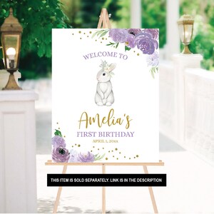 May include: A white welcome sign with purple flowers and gold glitter. The sign says "Welcome to Amelia's First Birthday April 1, 20xx".