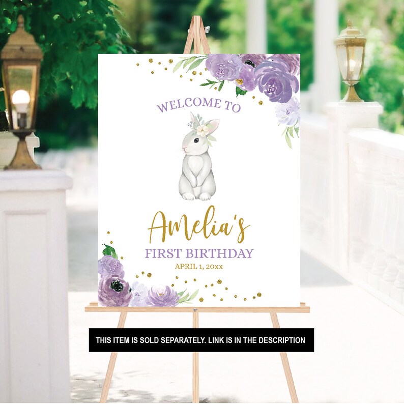 May include: A white welcome sign with purple flowers and gold glitter. The sign says "Welcome to Amelia's First Birthday April 1, 20xx".