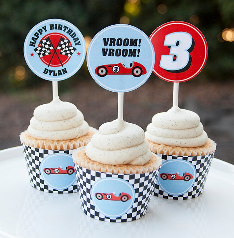 May include: Three cupcakes decorated with racing car themed toppers. The cupcakes have white frosting and are topped with a checkered cupcake liner. The toppers are round and feature a red race car, a checkered flag, and the number 3.