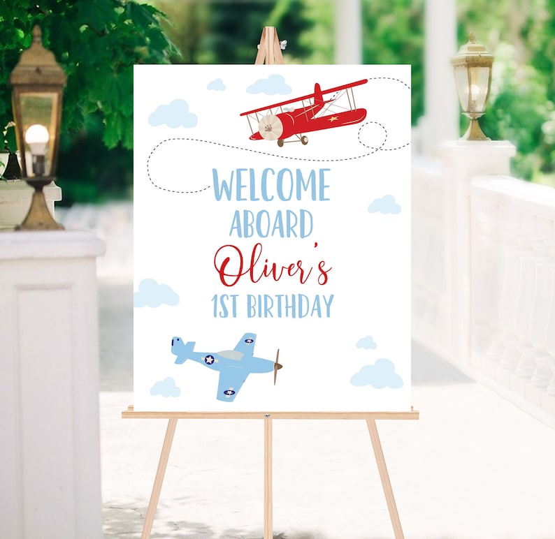May include: Welcome aboard Oliver's 1st birthday sign with a red and blue airplane illustration. The sign is on a wooden easel.