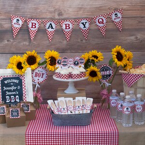 Może przedstawiać: St&oacute;ł na baby shower z czerwono-białym obrusem w kratkę. Na stole znajdują się babeczki, butelki z wodą i przekąski. St&oacute;ł jest ozdobiony słonecznikami i tabliczką z napisem "Witamy na Jennifer & Jason's BBQ".